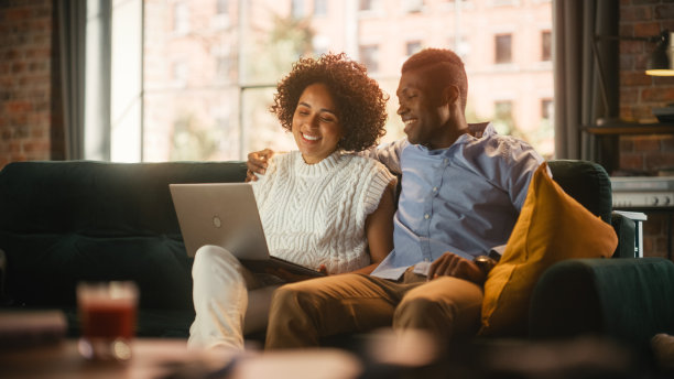 可爱的黑人情侣使用笔记本电脑，网上购物，使用社交媒体。男朋友和女朋友在舒适的公寓里坐在沙发上开心地笑。女性赋权。图片下载