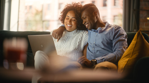 温柔的黑人夫妇使用笔记本电脑的肖像，坐在舒适的时尚公寓的沙发上。男女朋友在网上购物或看搞笑视频。图片下载