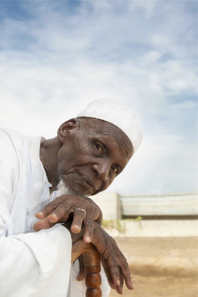 非洲老人坐在自家屋前，看着镜头，表情严肃，庆祝服装和帽子，八十岁高龄，照片图片下载