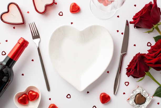 情人节的概念。顶部视图照片心形盘子刀叉茶托与糖果酒瓶玻璃蜡烛花束红玫瑰和纸屑在孤立的白色背景与复制空间图片下载