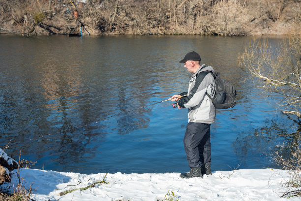 初春，一个渔夫背着背包，拿着鱼竿在白雪覆盖的河边捕鱼图片下载