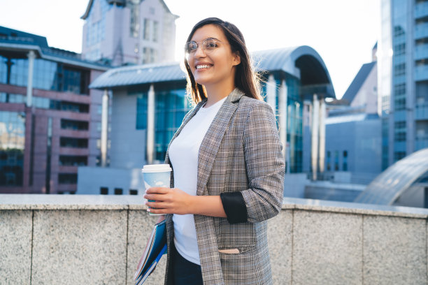 年轻的白人妇女穿着时髦的服装和眼镜站在城市设置在工作休息时享受阳光明媚的日子，时髦的女孩学生看着相机享受咖啡和休闲图片下载