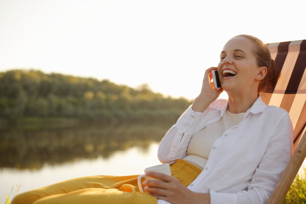非常快乐的美丽女人坐在河边的躺椅上，用智能手机聊天，谈笑风生，谈笑风生。图片下载