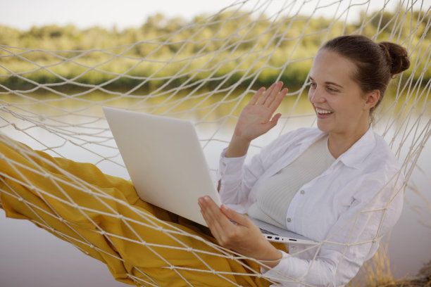 可爱的快乐积极棕色头发的女人的肖像，笔记本电脑在吊床上，与朋友进行视频通话或工作视频会议，挥手，打招呼。图片下载