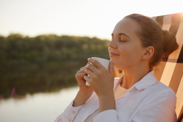 侧视图肖像快乐轻松平静的年轻女子穿着白衬衫放松在河边的躺椅上，手里拿着杯子，闻着令人愉快的咖啡香气，享受。图片下载