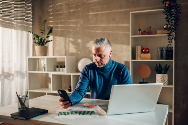 灰色头发的中年男子在家庭办公室工作，使用智能手机图片下载