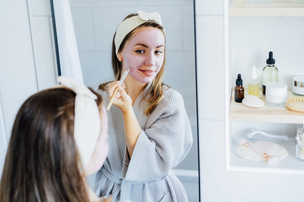 年轻女子在浴袍看着镜子和应用天然美容粘土面膜在她的脸在浴室。青少年在家护肤的美容程序。美容保健。有选择性的重点。图片下载