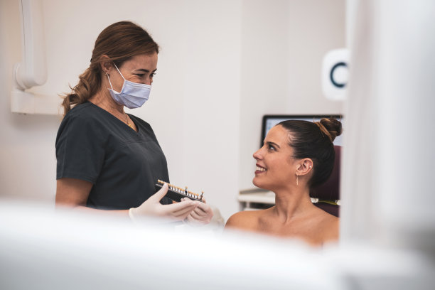牙科诊所里一位长着天然洁白牙齿的迷人年轻女子。一名女牙医展示了一个牙齿调色板，并互相看着比较颜色。白的牙齿。图片下载