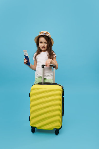 全身上下，小女孩穿着夏天的衣服，拿着机票，站在一个黄色的行李箱旁，孤立的蓝色背景图片下载