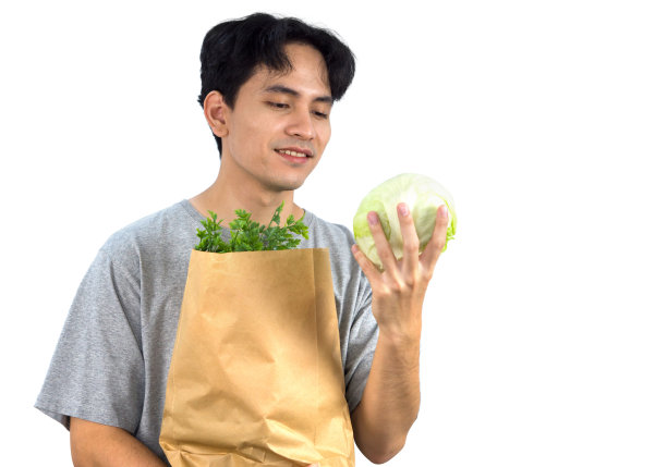 年轻的亚洲男子穿着便装，微笑着站在那里，手里拿着新鲜的生菜，手里提着装满蔬菜的纸袋。人像在白色背景与工作室灯光。图片下载