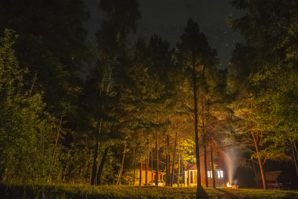 夜晚星空下，黑暗森林旁的房子的美丽景色。房子里的灯光为构图增添了光彩。夜间拍摄采用长曝光，选择性软对焦。图片下载