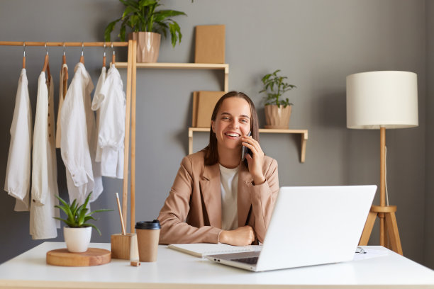 照片是穿着米色夹克的漂亮的白人女性设计师，在办公室里摆姿势，看着相机，笑得很开心。，与业务伙伴用智能手机通话。图片下载