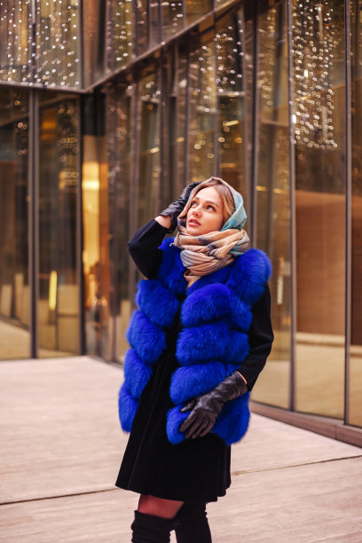 时尚漂亮的女人在蓝色毛皮背心和头巾在城区摆姿势，看向别处。时尚可爱的年轻女士走在户外。时尚风格概念。复制空白的广告文字图片下载