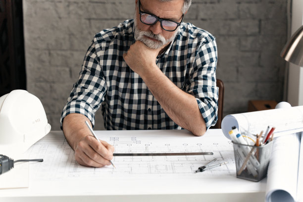 成熟自信，面带微笑的建筑师，穿着休闲装，在现代明亮的办公室里与蓝图文件和图纸一起工作。图片下载