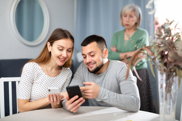 丈夫和妻子互相展示智能手机上的照片，一位不满的老年母亲站在他们身后图片下载