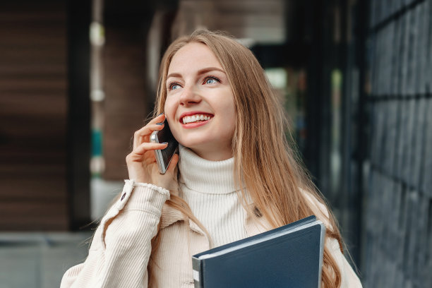金发学生女孩讲着手机，手里拿着笔记本文件夹，微笑着，看着大学大楼背景的一边。暗的背景图片下载