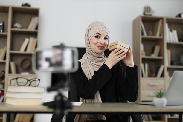 漂亮的穆斯林妇女在便装和头巾录制视频的相机，而打开礼盒。图片下载