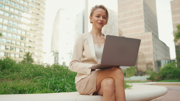 身穿米色西装的金发女商人正在外面使用笔记本电脑，脸上带着微笑图片下载