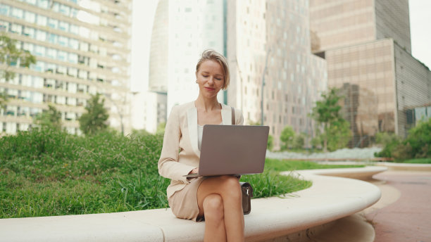 身穿米色西装的金发女商人正在外面使用笔记本电脑，脸上带着微笑图片下载