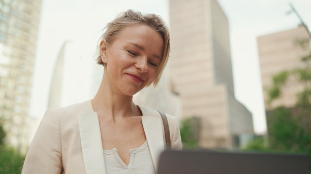身穿米色西装的金发女商人正在外面使用笔记本电脑，脸上带着微笑图片下载