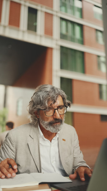 戴着眼镜留着胡子的成熟商人，坐在户外咖啡馆里，用笔记本电脑做笔记。成功的男人坐在桌子在咖啡馆在自然移动办公室自由职业者图片下载