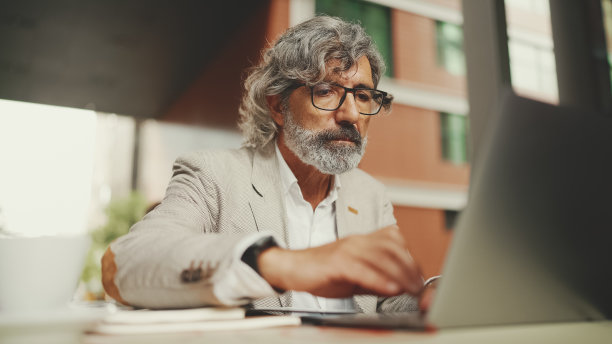 戴着眼镜留着胡子的成熟商人，坐在户外咖啡馆里，用笔记本电脑做笔记。成功的男人坐在桌子在咖啡馆在自然移动办公室自由职业者图片下载