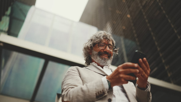 一位戴着眼镜、留着胡子的成熟商人站在商业中心背景的街道上。中年经理发短信，滚动点击到智能手机。交易带来的快乐图片下载