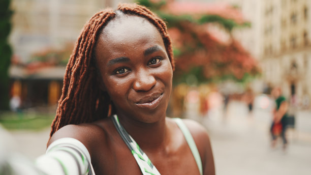 阴天，微笑着美丽的女人与非洲辫子穿着上衣站在外面的街道上，使用手机。时尚女孩自拍照片图片下载