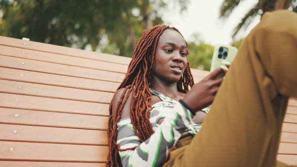 微笑的华丽女子与非洲辫子穿着在顶部是使用手机，而坐在长凳上。时髦的女孩看照片，视频在她的智能手机图片下载