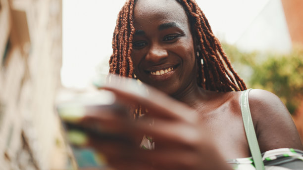 漂亮的女人与非洲辫子穿着上衣站在外面的街道上，使用手机。时尚女孩在智能手机上写信息图片下载