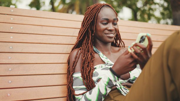 微笑的华丽女子与非洲辫子穿着在顶部是使用手机，而坐在长凳上。时髦的女孩看照片，视频在她的智能手机图片下载