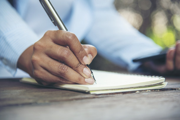 男人用手写笔记本、日记、咖啡杯和智能手机放在木头桌子上。近距离男子的手使用笔坐在木桌户外生活方式。男人写计划笔记、日记、办公室议程图片下载