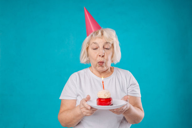 美丽快乐的高级妇女穿着白色t恤孤立地站在蓝色背景，拿着生日蛋糕和蜡烛在一个盘子上图片下载