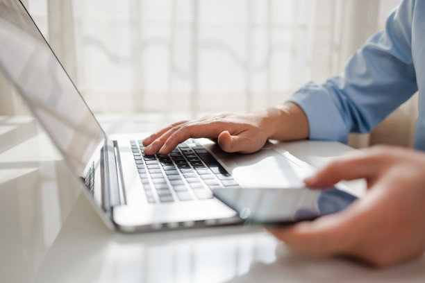 男性一边用智能手机，一边在笔记本电脑键盘上打字图片下载