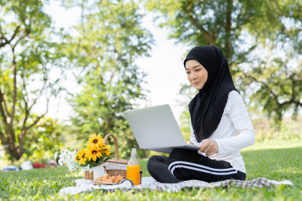 亚洲年轻的穆斯林妇女戴着黑头巾和希贾布裙子坐在公园里用笔记本电脑工作图片下载