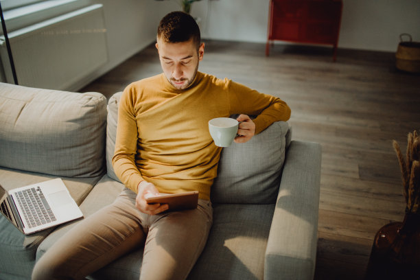 高角视角，一位男性企业家端着杯子坐在沙发上，用着平板电脑图片下载