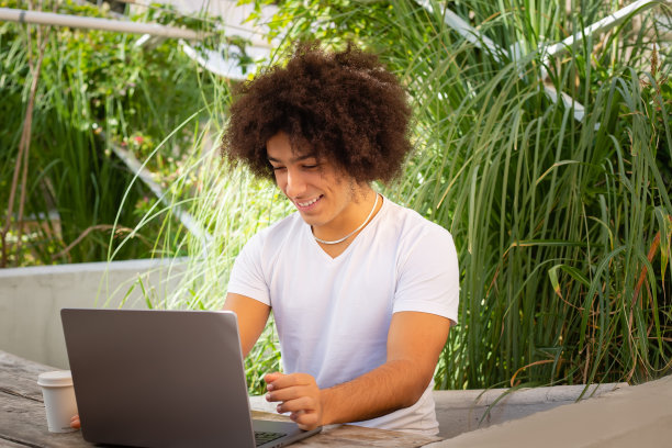 年轻的非裔意大利人微笑着在户外的绿色区域用笔记本电脑工作。自由的绿色开放空间是工作、商务、学习、自由职业的自然场所图片下载