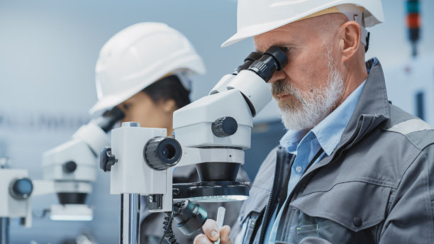 中年工业科学工程师在电子工厂设施的办公桌上工作，使用显微镜研究小型制造生产部件和细节。图片下载