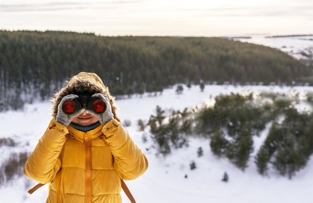 身穿黄色衣服的年轻女子微笑着用双筒望远镜在冰雪覆盖的河面上看鸟，在冬天的森林里观鸟，动物学，生态学研究自然，观察动物鸟类学复制空间图片下载