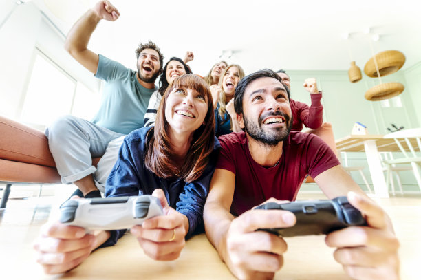 一群朋友在家里一起玩电子游戏，玩得很开心-男人和女人互相挑战，而朋友们坐在沙发上欢呼-友谊和游戏生活方式的概念图片下载