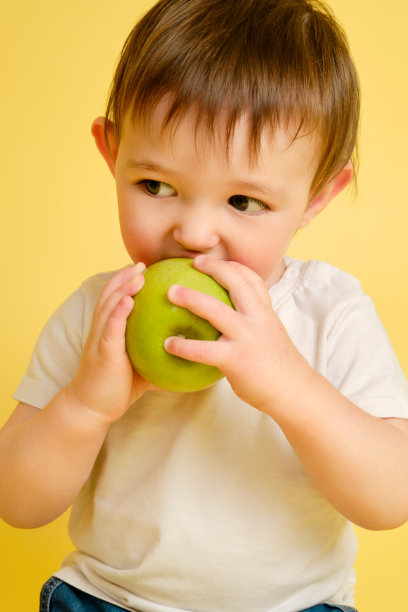 快乐的学步婴儿吃绿苹果工作室黄色背景。一个穿着白色t恤和蓝色牛仔裤，抱着一个大绿苹果的孩子。一岁零四个月的孩子图片下载