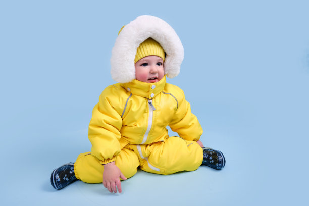 快乐的蹒跚学步的婴儿在冬天的衣服雪地服在工作室蓝色的背景。一个穿着带兜帽的温暖黄色连体衣的孩子。一岁五个月大的孩子图片下载