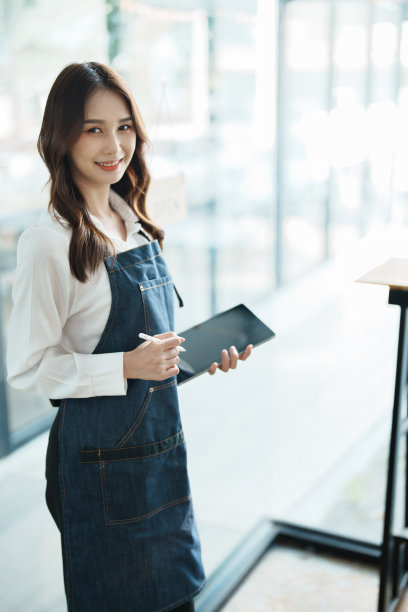 在咖啡馆前，穿着围裙，拿着平板电脑的快乐亚洲妇女的肖像。图片下载
