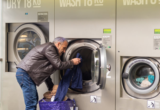 高级男性，穿着牛仔裤和棕色夹克，使用自助洗衣，插入脏衣服并使用卡支付，横向图像，复制空间图片下载