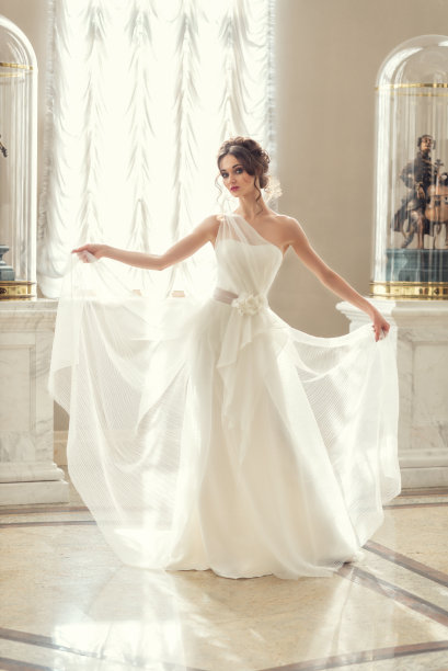 年轻漂亮的棕色头发的女人。身着豪华婚纱的新娘站在窗外的阳光下。婚礼发型和妆容。为美容院拍照图片下载