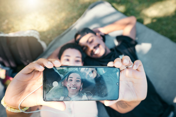 情侣在公园里，用手机自拍，躺在毯子上放松浪漫的野餐，共度美好时光。爱，浪漫，男人和女人在草地上，用智能手机屏幕上的照片和微笑。图片下载