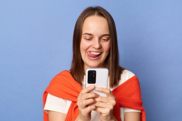 室内照片，正面女性穿着白色t恤和橙色毛衣，系在肩膀上，站在蓝色背景上，用手机，快乐地微笑，吐舌头。图片下载