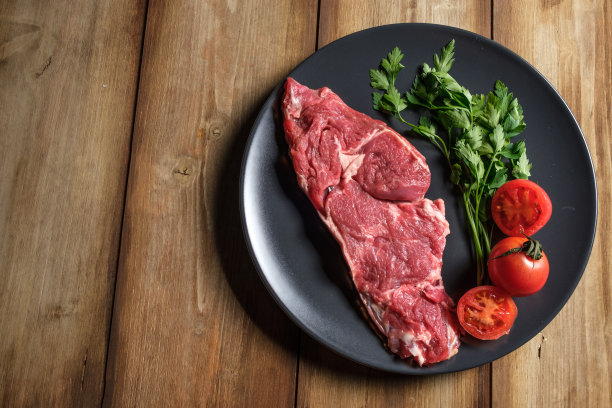 俯视图一块生牛肉与绿色蔬菜和西红柿上一个黑色的盘子图片下载