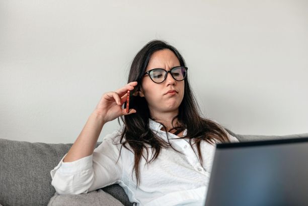 特写:一位戴眼镜的年轻女子坐在沙发上一边看笔记本电脑一边打电话图片下载
