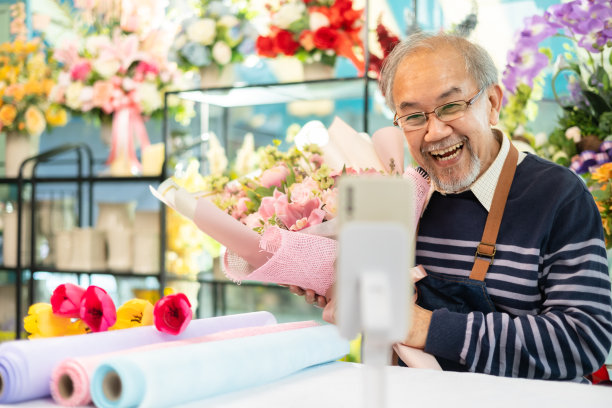 年长的高级男子在花店用笔记本电脑在智能手机上说话。成年男性小企业主在花店使用笔记本电脑和看着相机的肖像，图片下载